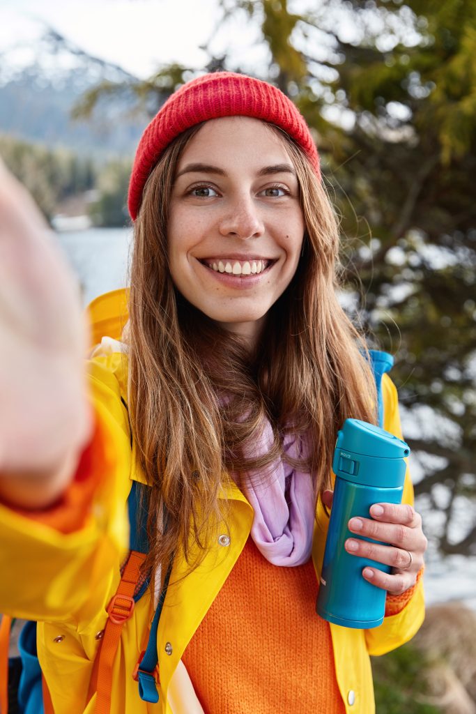 people, leisure and travelling concept. happy young european woman has toothy smile, relaxes on nature, makes selfie portrait as strolls on coniferous wood, holds thermos bottle of hot drink